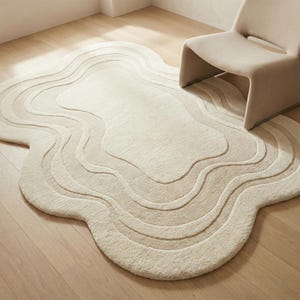 May include: A cream-coloured, abstract-shaped rug with a layered, wavy design. The rug is placed on a light-coloured wooden floor, with a matching chair in the background. The rug's design creates a sense of depth and texture.