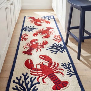May include: A cream-coloured rectangular rug with red lobster and navy blue coral designs. The rug has a navy blue border and is placed on a wooden floor. A navy blue stool is visible in the background.