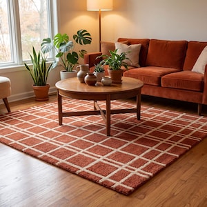 Pode incluir: Uma sala de estar com um tapete cor de ferrugem com um padrão de grade creme. Uma mesa de centro redonda de madeira exibe vasos e plantas em vasos. Um sofá cor de ferrugem e uma luminária de chão criam um ambiente acolhedor.