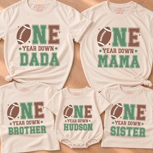 May include: A collection of five cream-colored shirts and one baby bodysuit featuring a football graphic and the text ONE YEAR DOWN with DADA, MAMA, BROTHER, HUDSON, or SISTER printed in green and brown block lettering on the front of each garment.