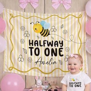 May include: A baby wearing a white onesie with the text "Halfway to One" and a bee graphic. A yellow and white striped backdrop with a bee illustration and the text "Halfway to One Amelia" is behind the baby. Pink balloons and bows decorate the scene.