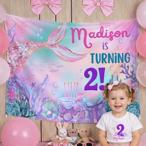 May include: A young girl smiles in front of a mermaid-themed birthday backdrop. The backdrop reads "Madison is turning 2!" and features a mermaid tail. Pink balloons and bows decorate the scene.