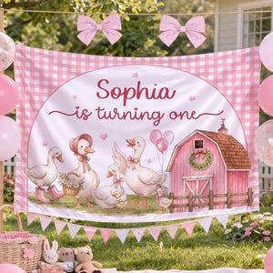 May include: A pink and white gingham backdrop featuring the text 'Sophia is turning one' with illustrations of white geese, a pink barn, and pink balloons. The banner is decorated with two large pink bows at the top and a pink triangular bunting at the bottom.