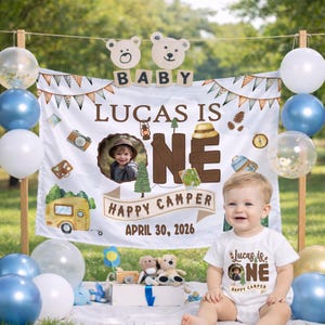May include: A baby's first birthday celebration with a "Happy Camper" theme. The backdrop features a banner with the text "Lucas is One" and a photo. The baby wears a matching onesie. Balloons in blue, white, and gold decorate the scene.