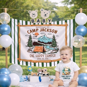 May include: A baby sits in front of a banner that says "Welcome to Camp Jackson" with a camping scene illustration. The baby wears a white onesie with a matching camping graphic. Balloons in blue, white, and gold decorate the scene.