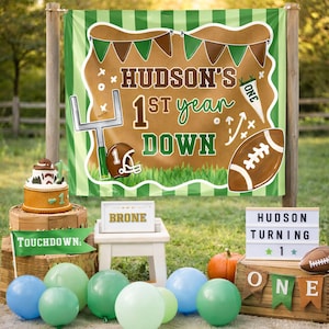 May include: A festive football-themed birthday party setup. A banner reads "HUDSON'S 1st year DOWN" with football graphics. Balloons, a cake, and a sign that says "HUDSON TURNING 1" complete the scene. A small pumpkin is also present.