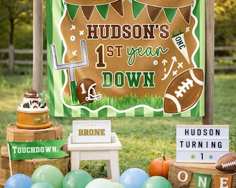 Pode incluir: Decoração de festa de aniversário com tema de futebol. Uma faixa diz "HUDSON'S 1st year DOWN" com gráficos de futebol. Balões, um bolo e uma placa que diz "HUDSON TURNING 1" completam a cena. Uma pequena abóbora também está presente.