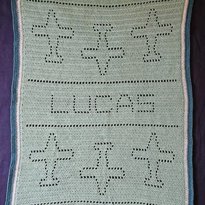May include: A crocheted blanket in a soft green hue, featuring a repeating airplane design and the name "LUCAS" in the center. The blanket has a teal border with white and teal accents, perfect for a baby or child.