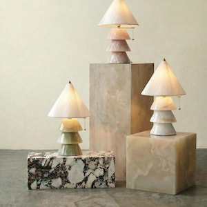 Könnte beinhalten: Drei dekorative Tischlampen mit konischen Schirmen und gestapelten Sockeln in Weiß, Rosa und Grün. Die Lampen stehen auf Steinsockeln mit Marmor- und Massivoberflächen und schaffen eine moderne, künstlerische Präsentation.