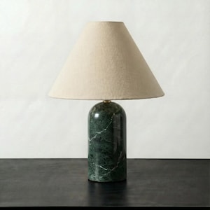 May include: A green marble table lamp with a beige lampshade. The lamp has a cylindrical base with white veining and a conical shade. The lamp is on a black surface.