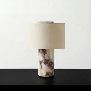 Könnte beinhalten: Zylindrische Tischlampe mit cremefarbenem Lampenschirm. Der Sockel besteht aus weißem Marmor mit lila Aderung. Die Lampe steht auf einer schwarzen Oberfläche vor weißem Hintergrund.