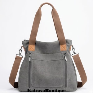 Puede incluir: Bolso tote de lona gris con correas y detalles marrones. Cuenta con un bolsillo frontal y dos compartimentos con cremallera. Incluye una correa de hombro desmontable y el texto "Kairava Boutique" en la parte inferior.