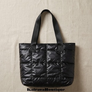 May include: Black quilted tote bag with black handles. The bag has a rectangular shape with a quilted pattern. The handles are made of a sturdy black material. The bag is sitting on a neutral background. The text "KairavaBoutique" is at the bottom.