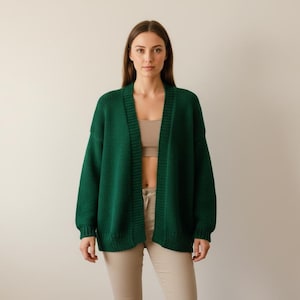 May include: A dark green, open-front cardigan sweater. The sweater has long sleeves and a relaxed fit. The model is wearing a beige top and khaki pants. The background is a solid, neutral color.