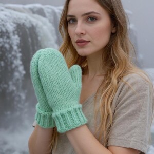 Op de afbeelding: Een paar mintgroene gebreide wanten. De wanten hebben een geribbelde manchet en een textuurgebreid patroon. De achtergrond toont een waterval en een persoon die een neutraal gekleurd shirt draagt.