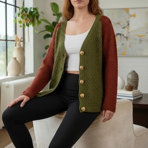 May include: A woman wearing a green and rust-colored cardigan with gold buttons. The cardigan is paired with a white top and black leggings. The cardigan has a textured knit pattern.