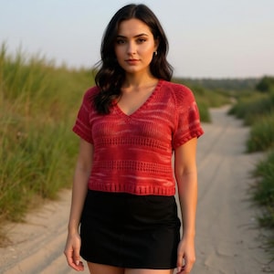 May include: A red short-sleeved knit top with a V-neck. The top has a horizontal striped pattern and is paired with a black skirt. The image is taken outdoors on a dirt path with green grass in the background.