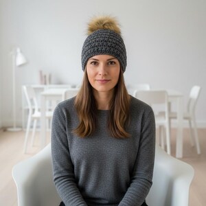 May include: A gray knit beanie with a faux fur pom-pom. The hat is worn by a person wearing a gray long-sleeve sweater. The background is a bright room with white furniture.