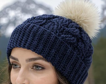 May include: A navy blue knit hat with a textured pattern and a large, fluffy, cream-colored pom-pom. The hat is made of a thick, warm yarn, perfect for cold weather. The hat is a winter accessory.