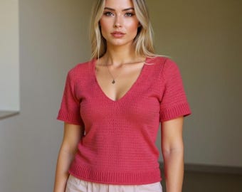 Puede incluir: Una camiseta de punto de color coral de manga corta con cuello en V. La parte superior tiene un diseño de tejido abierto texturizado y puños y dobladillo acanalados. La modelo lleva un delicado collar. La parte superior se combina con pantalones de color claro.