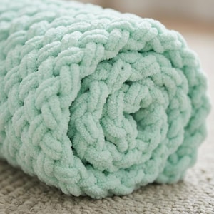May include: A rolled-up, chunky knit blanket in a soft, pastel green color. The blanket is made of thick, plush yarn, creating a textured surface. The close-up shot highlights the cozy, inviting nature of the blanket, perfect for home decor.