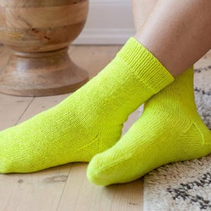 May include: A pair of vibrant neon yellow socks. The socks are knitted with a ribbed cuff. The socks are on a person's feet, with one foot extended and the other bent. The background includes a wooden stool and a patterned rug.