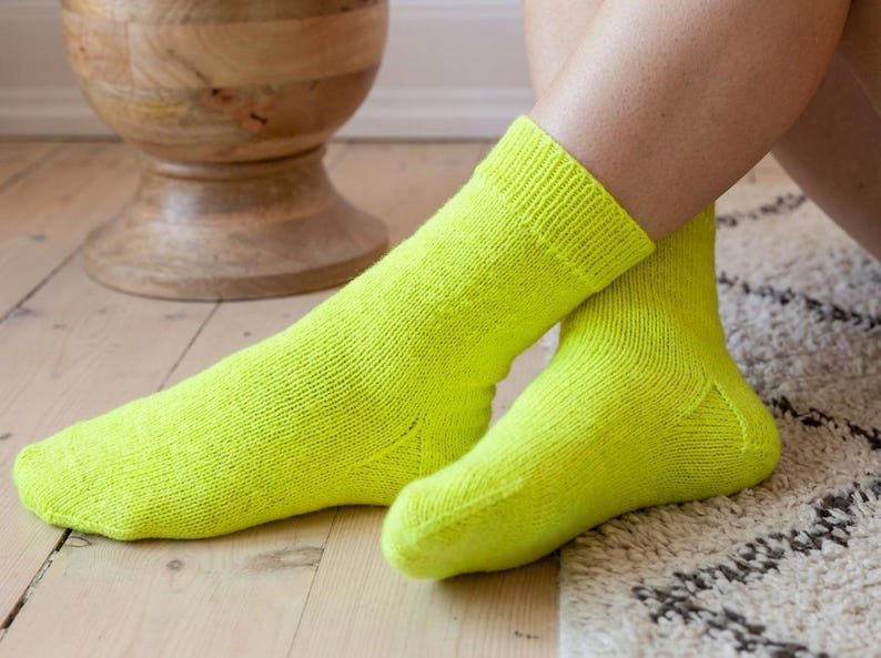 May include: A pair of vibrant neon yellow socks. The socks are knitted with a ribbed cuff. The socks are on a person's feet, with one foot extended and the other bent. The background includes a wooden stool and a patterned rug.