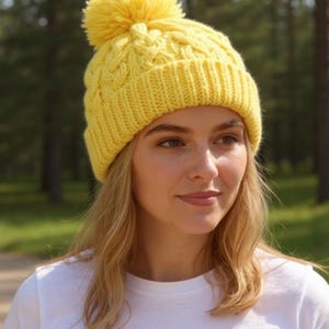 May include: A vibrant yellow cable-knit beanie hat with a fluffy pom-pom. The hat is made of a soft, textured yarn. The person in the image is wearing a white t-shirt. The background is a blurred view of a forest.