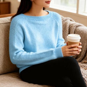 Op de afbeelding: Een lichtblauwe trui met geribbelde details op de mouwen en manchetten. De trui heeft een ronde hals en is gemaakt van een zacht, mogelijk gebreid materiaal. De persoon houdt een papieren koffiebeker vast.