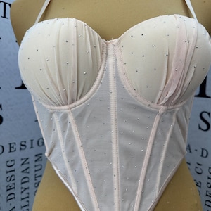 May include: A beige corset top featuring a sheer mesh overlay with scattered silver rhinestones. The garment has structured cups with gathered fabric detailing, thin shoulder straps, and a pointed hemline, displayed on a mannequin torso.
