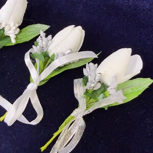 Op de afbeelding: Drie witte tulp corsages met groene bladeren, zilveren glitteraccenten en zilveren linten. Elk bloemstuk is opgebouwd uit een enkele witte tulpenknop, groen blad en decoratieve zilveren elementen.