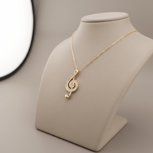 May include: A gold-toned necklace featuring a treble clef pendant. The pendant is encrusted with small, clear, sparkling stones along its curved shape. The necklace hangs from a delicate gold-toned chain, creating a refined and elegant piece of jewelry.