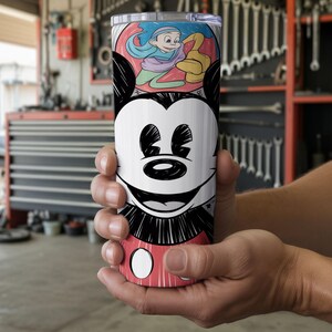 May include: A stainless steel tumbler featuring a black and white Mickey Mouse face design. The tumbler also has a colorful illustration of a cartoon character. The tumbler is held in a person's hands.
