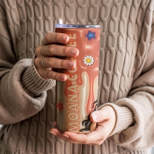 K&ouml;nnte beinhalten: Ein Edelstahlbecher mit Deckel und Strohhalm, mit dem Text "MOANA COFFEE" in Orange. Das Design umfasst einen blauen Stern, eine G&auml;nsebl&uuml;mchenblume und einen tropfenden Kaffeeeffekt in Creme- und Braunt&ouml;nen.