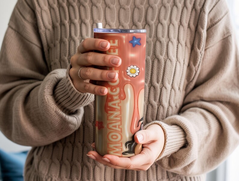 K&ouml;nnte beinhalten: Ein Edelstahlbecher mit Deckel und Strohhalm, mit dem Text "MOANA COFFEE" in Orange. Das Design umfasst einen blauen Stern, eine G&auml;nsebl&uuml;mchenblume und einen tropfenden Kaffeeeffekt in Creme- und Braunt&ouml;nen.
