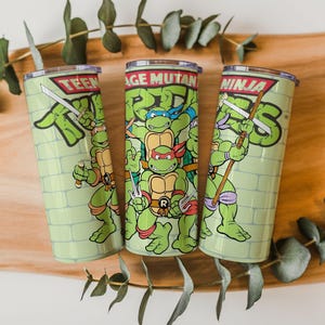 May include: Three green stainless steel tumblers featuring the Teenage Mutant Ninja Turtles. Each tumbler has a different Ninja Turtle character with their weapon. The tumblers have a light green brick wall background and the words "Teenage Mutant Ninja Turtles".