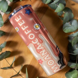 K&ouml;nnte beinhalten: Ein Edelstahlbecher mit rosa und wei&szlig;em Design. Der Becher zeigt die Worte "MOANA COFFEE" in einer verspielten Schriftart sowie G&auml;nsebl&uuml;mchen- und Sterngrafiken. Der Becher wird auf einer Holzoberfl&auml;che mit gr&uuml;nem Laub gezeigt.