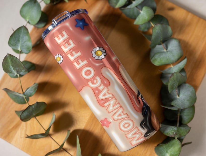 K&ouml;nnte beinhalten: Ein Edelstahlbecher mit rosa und wei&szlig;em Design. Der Becher zeigt die Worte "MOANA COFFEE" in einer verspielten Schriftart sowie G&auml;nsebl&uuml;mchen- und Sterngrafiken. Der Becher wird auf einer Holzoberfl&auml;che mit gr&uuml;nem Laub gezeigt.
