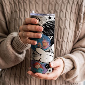 May include: A tall, insulated tumbler with a whimsical cartoon design. The tumbler features a white and black Mickey Mouse hand, with colorful illustrations of characters and scenes. The design includes a blue background with a lantern and a red-haired character.