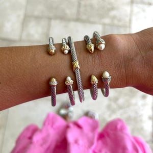 May include: A collection of silver and gold cable bracelets worn on the wrist. The bracelets feature twisted metal bands with gold end caps, some accented with small white pearls and others with clear sparkling stones. One bracelet has an X-shaped gold center detail.