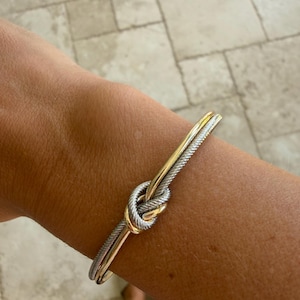 Könnte beinhalten: Ein zweifarbiger Armreif bestehend aus einem polierten goldfarbenen Metallband und einem strukturierten silberfarbenen Kabelband, die zu einem zentralen Knoten verschlungen sind. Der Armreif hat eine starre, offene Struktur am Handgelenk.