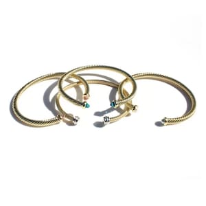 May include: Three gold-toned cable-style cuff bracelets with open ends. Each bracelet features decorative end caps in varying colors, including teal, white, and metallic tones, attached to the twisted metal bands that form a circular shape.