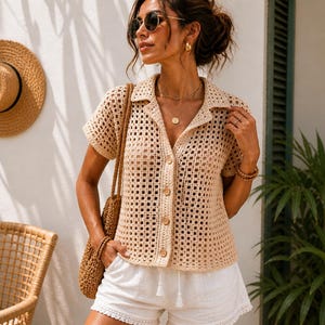May include: A beige crochet shirt with short sleeves and wooden buttons is worn with white shorts. A straw hat hangs on the wall, and a woven bag is slung over the shoulder. The outfit is accessorized with sunglasses and gold jewellery.