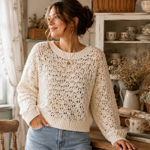 Può includere: Maglione traforato color crema con scollo tondo e maniche lunghe. Il maglione è abbinato a jeans blu. L'immagine è ambientata in una stanza accogliente e ben illuminata con un'estetica vintage.