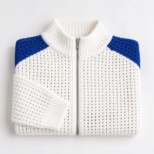 Puede incluir: Suéter blanco de punto con cremallera y detalles azules en los hombros. El suéter tiene un cuello alto y una cremallera plateada de largo completo. El punto tiene un diseño perforado y las mangas están dobladas. El suéter está doblado sobre una superficie blanca.