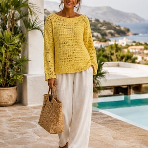 Può includere: Una donna indossa un maglione giallo all'uncinetto, pantaloni di lino bianchi e sandali marroni, con una borsa tote intrecciata. Il maglione ha maniche lunghe e scollatura a barchetta. Lo sfondo mostra una piscina, il mare e vegetazione.