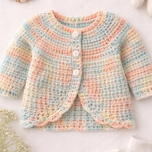 May include: A pastel-colored, crocheted baby cardigan with a scalloped edge. The soft, multi-colored yarn includes shades of pink, blue, and yellow. The cardigan has three white buttons and long sleeves. A wooden teething ring and a stuffed animal are in the background.