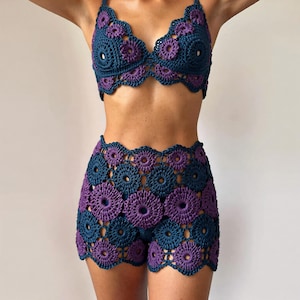 Op de afbeelding: Een tweedelige gehaakte set in blauw en paars met een bloemenpatroon. De set bestaat uit een bh-top en een high-waisted short, beide met geschulpte randen. Het haakwerk is gedetailleerd en ingewikkeld.