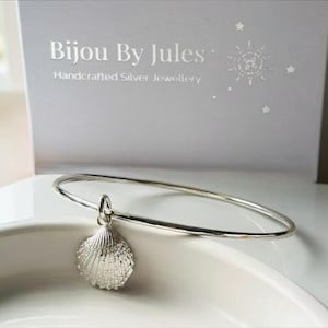 May include: A silver bangle bracelet featuring a textured seashell charm. The bracelet is positioned in front of a light grey display card with the text Bijou By Jules Handcrafted Silver Jewellery and a small star graphic printed in silver.