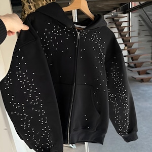 Puede incluir: Sudadera negra con cremallera y adornos blancos en forma de estrella en las mangas y la parte delantera. La sudadera con capucha tiene una capucha, una cremallera completa y un bolsillo delantero. La prenda está colgada en una percha de madera.